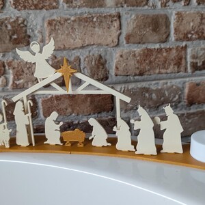 May include: A wooden nativity scene with an angel, star, and figures of Mary, Joseph, and the Three Wise Men. The scene is set on a gold-colored base, with two battery-operated LED candles on either end. The background is a brick wall.