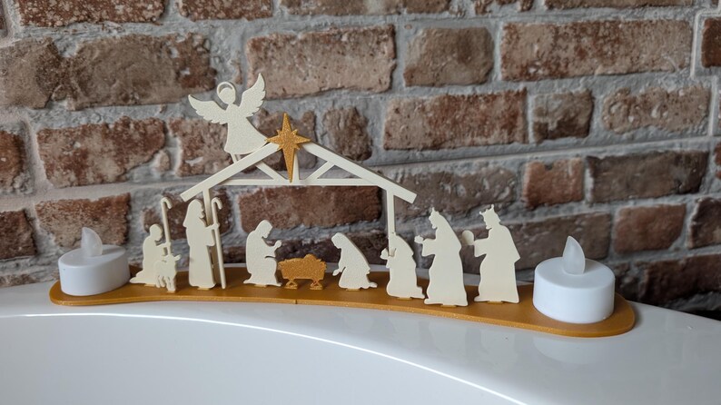 May include: A wooden nativity scene with an angel, star, and figures of Mary, Joseph, and the Three Wise Men. The scene is set on a gold-colored base, with two battery-operated LED candles on either end. The background is a brick wall.