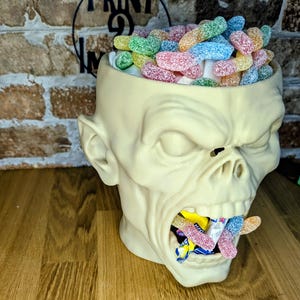 May include: A cream-colored zombie head-shaped container filled with colorful gummy candies. The container's mouth is also filled with candies. The background includes a brick wall and the text "PRINT 9 IN".