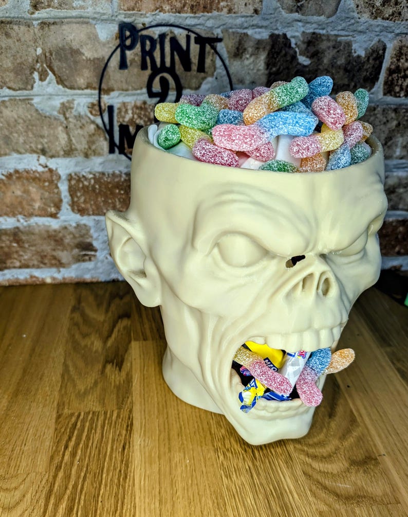 May include: A cream-colored zombie head-shaped container filled with colorful gummy candies. The container's mouth is also filled with candies. The background includes a brick wall and the text "PRINT 9 IN".