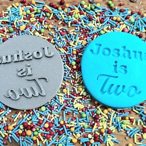 May include: Two circular cookie stamps, one gray and one blue, with the text "Joshua is Two" and "Joshua is out" in a script font. The stamps are surrounded by colorful sprinkles.