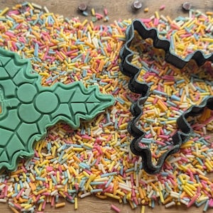 May include: Green holly leaf and black heart-shaped cookie cutters rest on a bed of colorful sprinkles. The holly leaf cutter has detailed leaf and berry designs. The sprinkles are a mix of yellow, pink, orange, and blue.