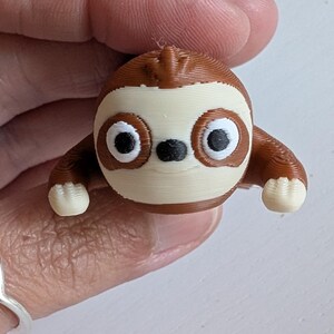 May include: A 3D-printed sloth figurine in shades of brown, cream, and black. The sloth has large, round eyes and small arms and feet. The figurine is held in a hand.