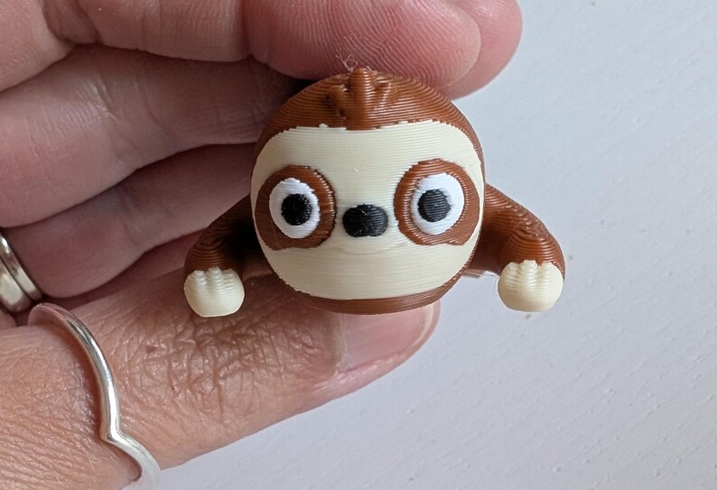 May include: A 3D-printed sloth figurine in shades of brown, cream, and black. The sloth has large, round eyes and small arms and feet. The figurine is held in a hand.