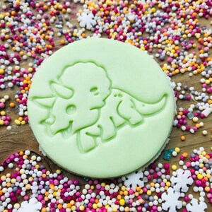 May include: A light green, circular cookie cutter featuring a detailed design of a triceratops dinosaur. The cutter is surrounded by colorful sprinkles, suggesting a baking or crafting theme. The design is embossed, ready to create a dinosaur-shaped treat.