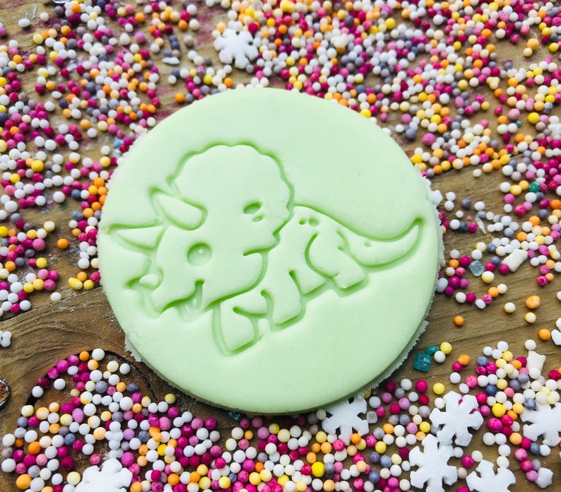 May include: A light green, circular cookie cutter featuring a detailed design of a triceratops dinosaur. The cutter is surrounded by colorful sprinkles, suggesting a baking or crafting theme. The design is embossed, ready to create a dinosaur-shaped treat.
