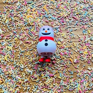 May include: A white snowman figurine with a red scarf and black eyes and buttons, surrounded by colorful sprinkles on a wooden surface.