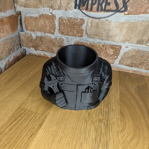 May include: A black planter shaped like a mechanic holding a wrench. The planter has a large opening for plants and is designed to resemble a mechanic's uniform, complete with pockets and details.