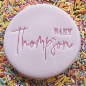 May include: A pink round cookie with the words "BABY Thompson" in pink lettering.