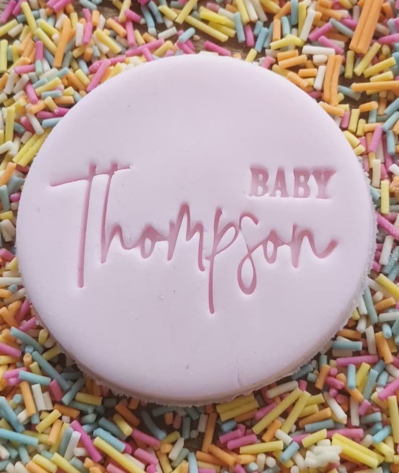 May include: A pink round cookie with the words "BABY Thompson" in pink lettering.