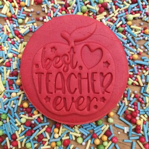 May include: A round red cookie with the words "best teacher ever" embossed on it, featuring an apple design with a heart and stars. It is surrounded by colourful sprinkles on a wooden surface.
