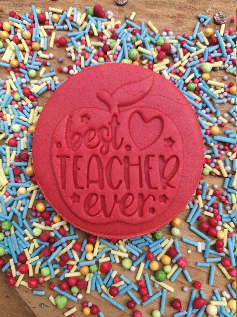 May include: A round red cookie with the words "best teacher ever" embossed on it, featuring an apple design with a heart and stars. It is surrounded by colourful sprinkles on a wooden surface.