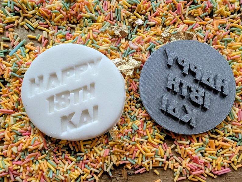 May include: A white, round edible cake decoration with the words "HAPPY 18TH KAI" embossed on it, next to a gray, round stamp with the same text in reverse. The items are on a bed of colorful sprinkles.