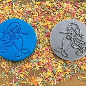 Puede incluir: Dos cortadores de galletas de silicona azul y gris con forma de sirenas con cabello largo y fluido. Los cortadores est&aacute;n sobre un fondo de chispas de colores.