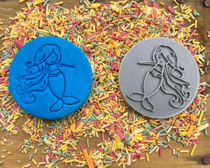 Puede incluir: Dos cortadores de galletas de silicona azul y gris con forma de sirenas con cabello largo y fluido. Los cortadores est&aacute;n sobre un fondo de chispas de colores.