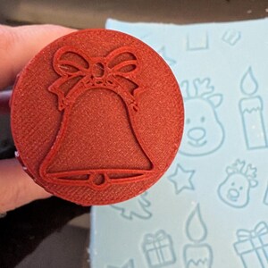 May include: A red, circular stamp with a bell design and a bow at the top. The stamp is held over a light blue surface with Christmas-themed patterns, including reindeer, candles, stars, and gifts. This stamp is likely for crafting or baking.