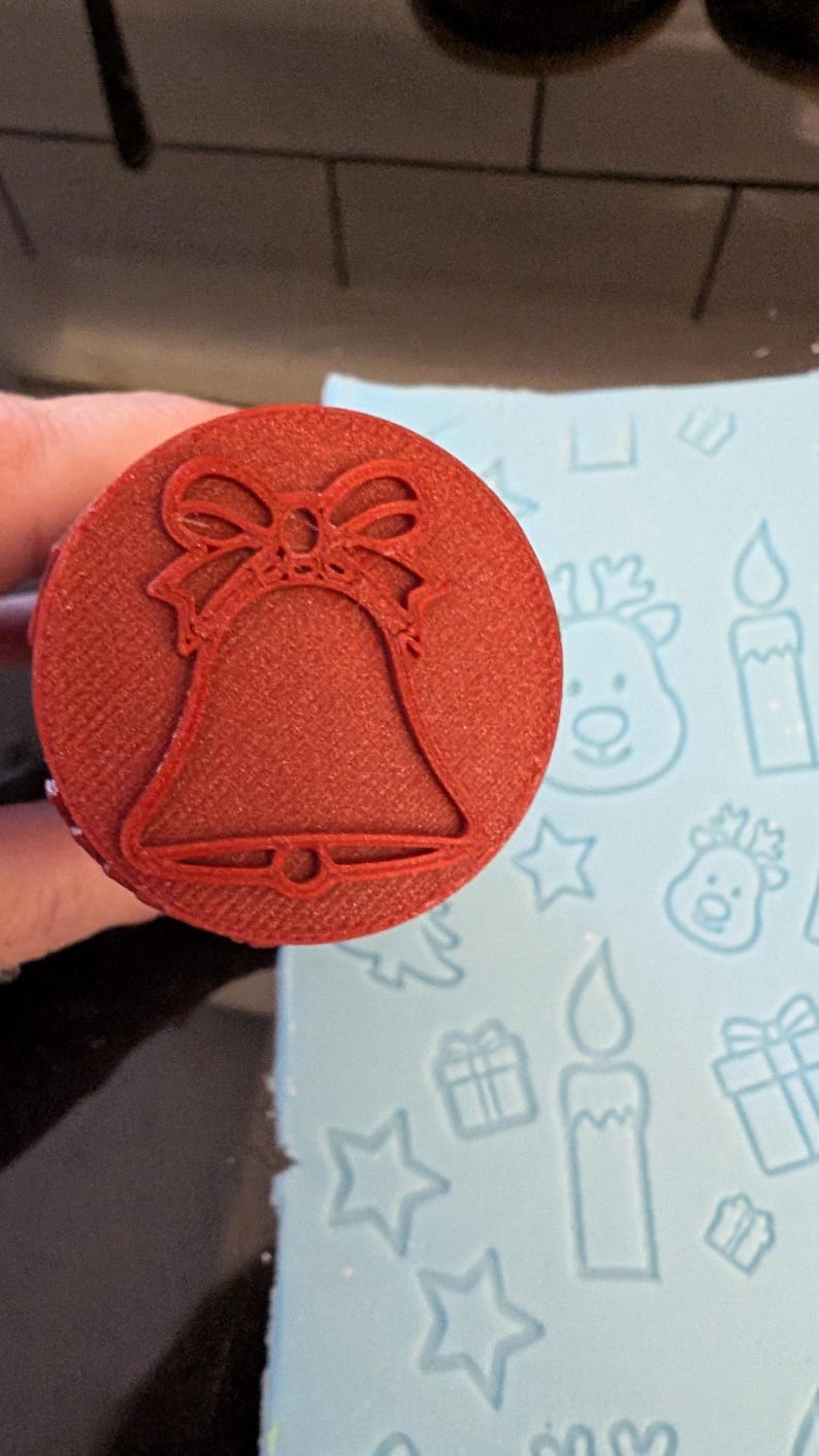 May include: A red, circular stamp with a bell design and a bow at the top. The stamp is held over a light blue surface with Christmas-themed patterns, including reindeer, candles, stars, and gifts. This stamp is likely for crafting or baking.