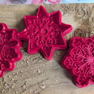 May include: Three red plastic snowflake-shaped cookie cutters. One is a six-pointed star, and the other two are circular with intricate snowflake designs. The cutters are on a weathered wooden surface.