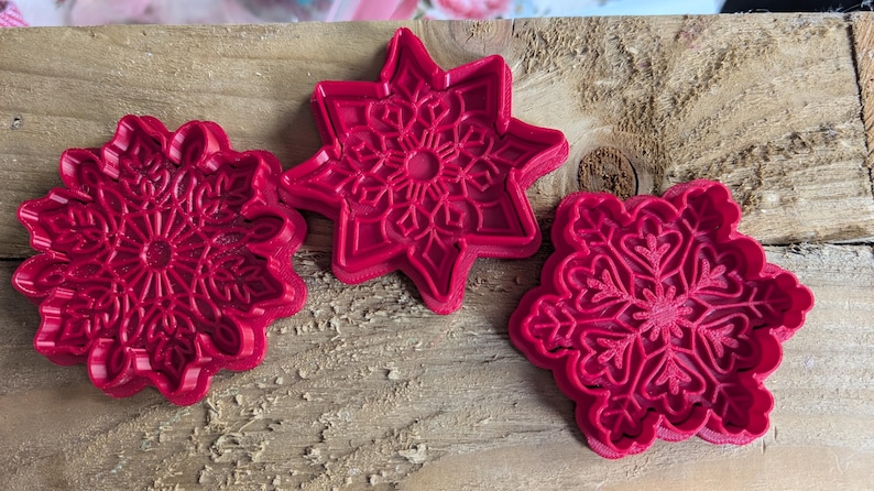 May include: Three red plastic snowflake-shaped cookie cutters. One is a six-pointed star, and the other two are circular with intricate snowflake designs. The cutters are on a weathered wooden surface.
