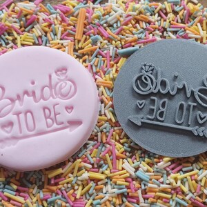 May include: Two round stamps with the text "Bride TO BE" and an arrow. One stamp is pink, the other is gray. The stamps are surrounded by colorful sprinkles, creating a festive and celebratory theme.
