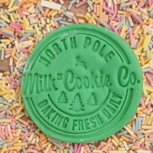Może przedstawiać: Zielona okrągła pieczątka do ciasteczek z tekstem "North Pole Milk & Cookie Co. Baking Fresh Daily" i trzema małymi ilustracjami sosen.