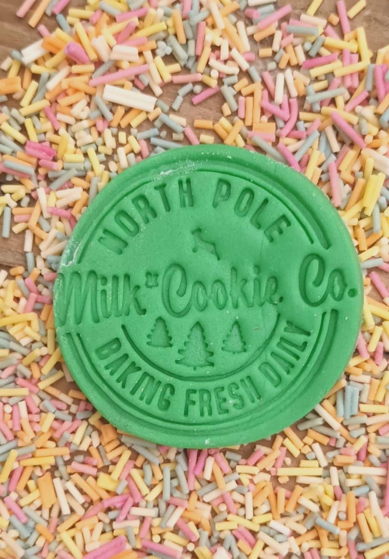 Może przedstawiać: Zielona okrągła pieczątka do ciasteczek z tekstem "North Pole Milk & Cookie Co. Baking Fresh Daily" i trzema małymi ilustracjami sosen.