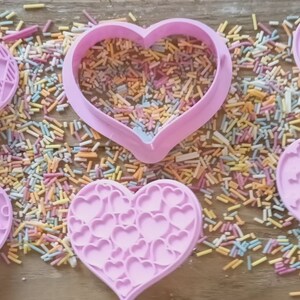 Puede incluir: Seis cortadores de galletas rosas en forma de coraz&oacute;n con varios dise&ntilde;os en relieve, incluyendo un rompecabezas y un patr&oacute;n de rosas. Los cortadores est&aacute;n rodeados de chispas de colores, sugiriendo un tema de reposter&iacute;a.