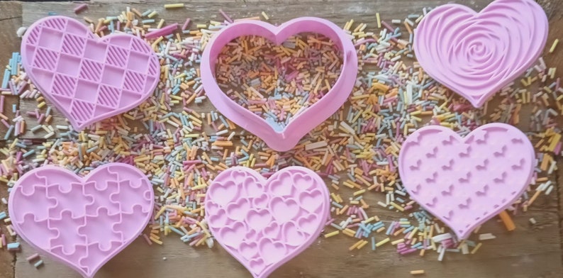 Puede incluir: Seis cortadores de galletas rosas en forma de coraz&oacute;n con varios dise&ntilde;os en relieve, incluyendo un rompecabezas y un patr&oacute;n de rosas. Los cortadores est&aacute;n rodeados de chispas de colores, sugiriendo un tema de reposter&iacute;a.