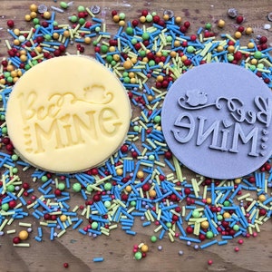 K&ouml;nnte beinhalten: Ein gelber, runder Keks mit den Worten "Bee Mine" gepr&auml;gt, neben einem grauen, runden Stempel mit dem gleichen Design. Die Artikel sind von bunten Streuseln auf einer Holzoberfl&auml;che umgeben.