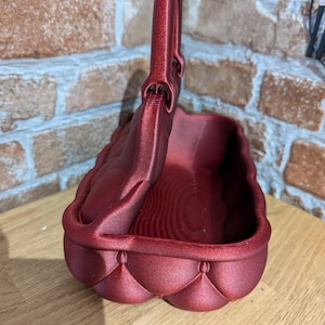 May include: A burgundy-colored basket with a handle. The basket has a quilted design on the sides and a textured interior. The handle is arched and attached to the top of the basket. The basket is sitting on a wooden surface.