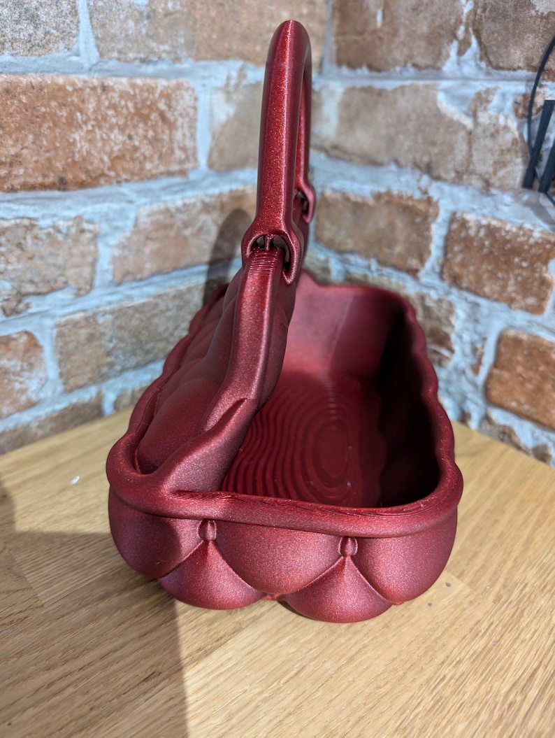 May include: A burgundy-colored basket with a handle. The basket has a quilted design on the sides and a textured interior. The handle is arched and attached to the top of the basket. The basket is sitting on a wooden surface.