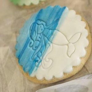 Puede incluir: Una galleta de az&uacute;car decorada con glaseado azul y blanco. El glaseado representa una sirena con el pelo largo y suelto.