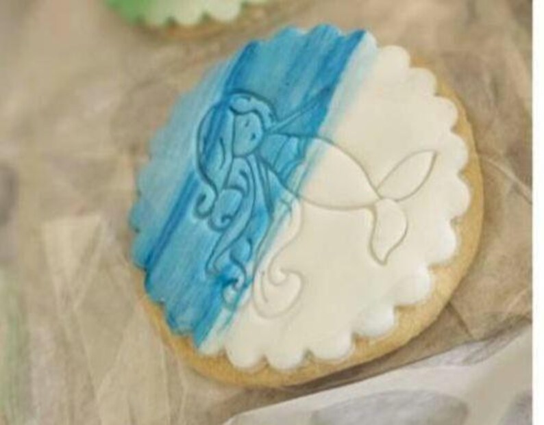 Puede incluir: Una galleta de az&uacute;car decorada con glaseado azul y blanco. El glaseado representa una sirena con el pelo largo y suelto.