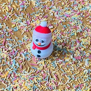 May include: A white snowman figurine with a red hat and scarf, surrounded by colorful sprinkles on a wooden surface.