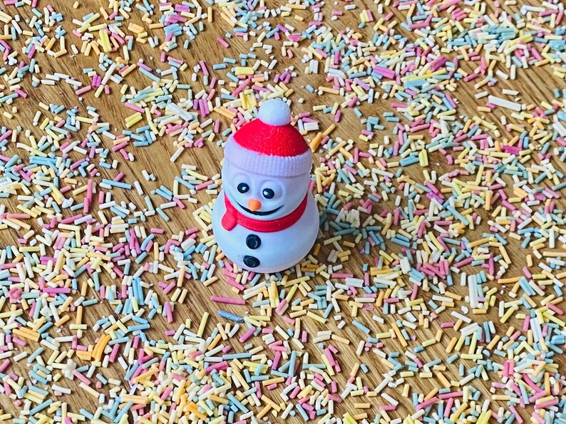 May include: A white snowman figurine with a red hat and scarf, surrounded by colorful sprinkles on a wooden surface.