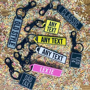 May include: A collection of personalized keychains with various designs and text. The keychains feature black metal rings and different colored backgrounds, including gray, yellow, white, pink, and black. The text on the keychains includes "OLD STYLE", "CLASSIC", "ANY TEXT", "LEX1E", "DAD TAX1", and "LEO 14".