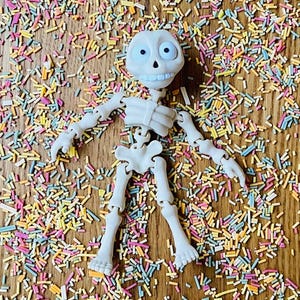 May include: A white plastic skeleton toy with movable joints, lying on a wooden surface covered in colorful sprinkles.