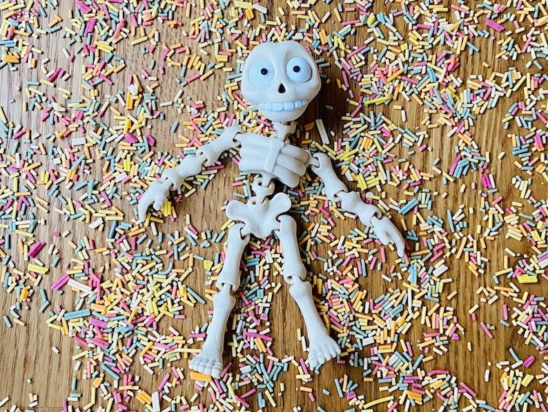 May include: A white plastic skeleton toy with movable joints, lying on a wooden surface covered in colorful sprinkles.
