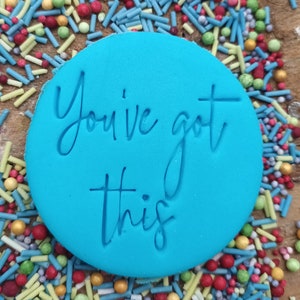 以下が含まれることがあります： 「You've got this」の文字が筆記体で刻まれた、明るい青色の丸いフォンダン。赤、緑、黄色、青のカラフルなスプリンクルで囲まれています。