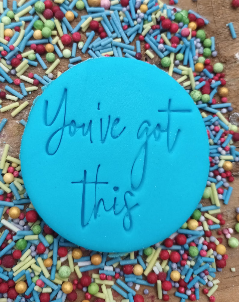 以下が含まれることがあります： 「You've got this」の文字が筆記体で刻まれた、明るい青色の丸いフォンダン。赤、緑、黄色、青のカラフルなスプリンクルで囲まれています。