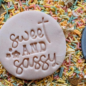 May include: A light pink, circular cookie stamp with the words "Sweet AND Sassy" carved into it, along with a cross and flower design. The stamp is surrounded by colorful sprinkles.