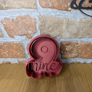 May include: A red plastic cookie cutter in the shape of the number nine with the word "nine" written below it. The cookie cutter is on a wooden surface in front of a brick wall.