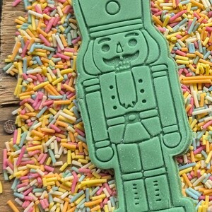 May include: A green nutcracker-shaped cookie cutter on a wooden surface, surrounded by colorful sprinkles. The cookie cutter has detailed features, including a hat, face, and uniform. The sprinkles are a mix of yellow, pink, blue, and white.