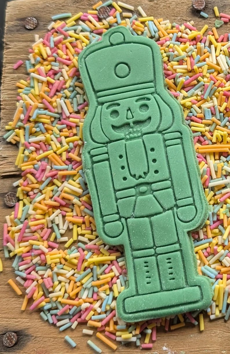 May include: A green nutcracker-shaped cookie cutter on a wooden surface, surrounded by colorful sprinkles. The cookie cutter has detailed features, including a hat, face, and uniform. The sprinkles are a mix of yellow, pink, blue, and white.