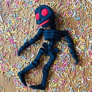 May include: A black articulated skeleton toy with a red skull lying on a surface covered in colorful sprinkles.