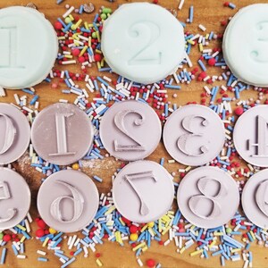 May include: A collection of circular number stamps, including numbers 0 through 9, and three light blue number-shaped candies. The stamps are gray and sit on a wooden surface scattered with colorful sprinkles.
