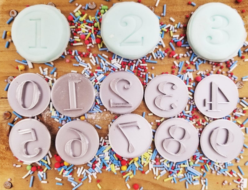 May include: A collection of circular number stamps, including numbers 0 through 9, and three light blue number-shaped candies. The stamps are gray and sit on a wooden surface scattered with colorful sprinkles.