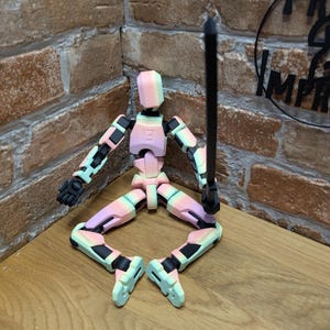 May include: A poseable wooden artist's mannequin in a seated position, painted with pastel colors. The figure holds a black rod and has articulated joints. The mannequin is on a wooden surface with a brick wall in the background.