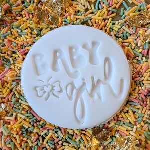 May include: A white circular fondant with the words "BABY girl" embossed on it, along with a bow and heart design. The fondant is surrounded by colorful sprinkles and gold leaf flakes.