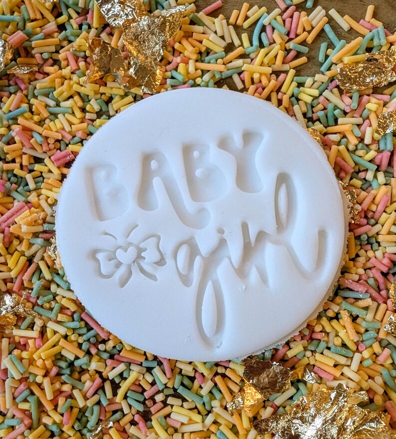May include: A white circular fondant with the words "BABY girl" embossed on it, along with a bow and heart design. The fondant is surrounded by colorful sprinkles and gold leaf flakes.